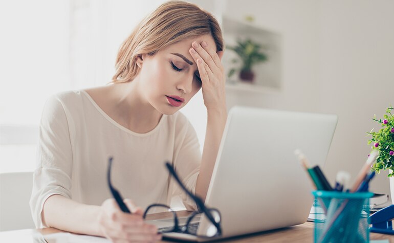 Eine Frau, die vor einem Laptop sitzt, schließt ihre Augen und greift sich an die Stirn